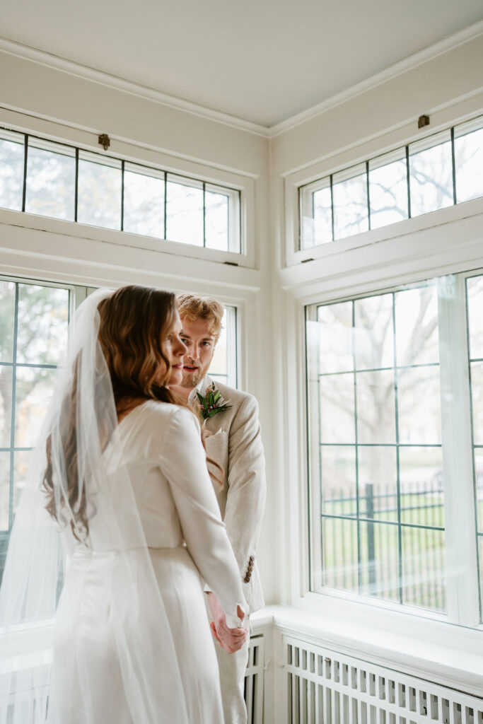 Bride and groom stand in the corner of a white room with natural lighting