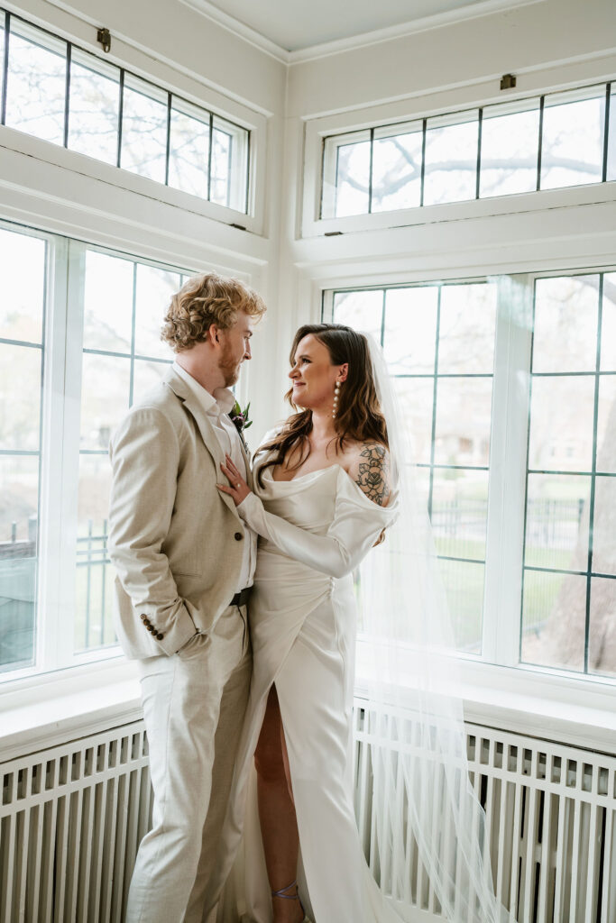 Bride and groom stand in the corner of a white room with natural lighting