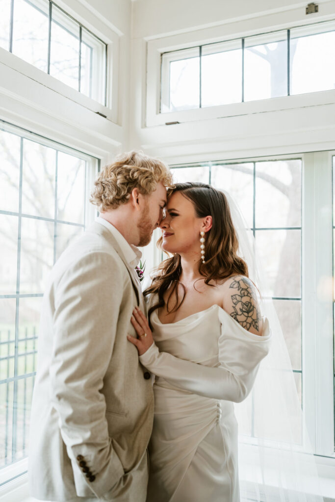 Bride and groom stand in the corner of a white room with natural lighting