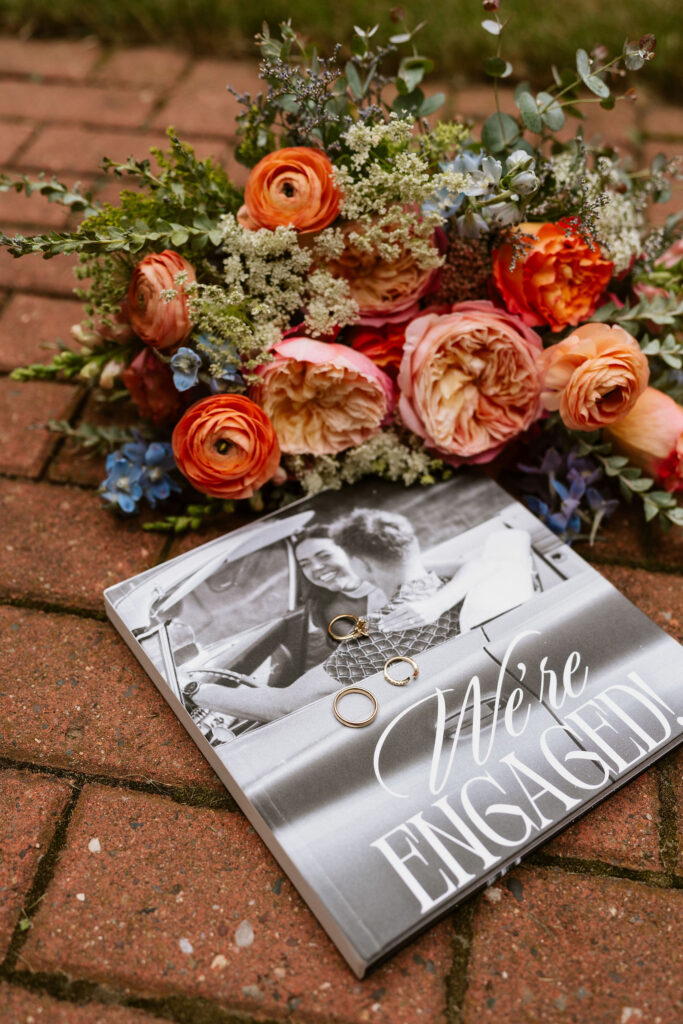 A colorful wedding bouquet lays on a brick walkway next to an engagement photo album