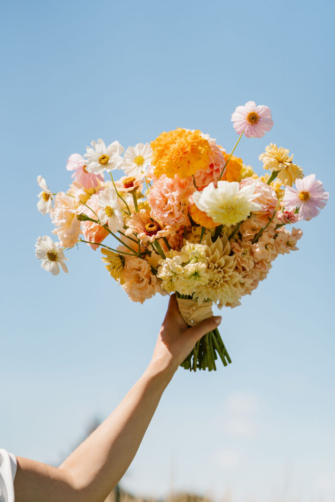 Bride holds up her colorful orange and yellow bouquet to the blue sky